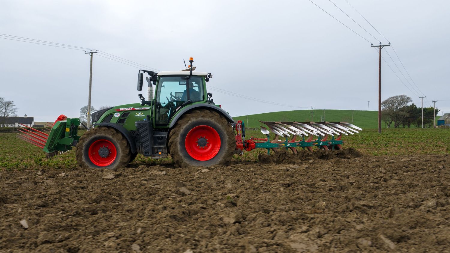 Green Fendt tractor with red wheels plowing a muddy field using a multi-furrow plow on a cloudy day.