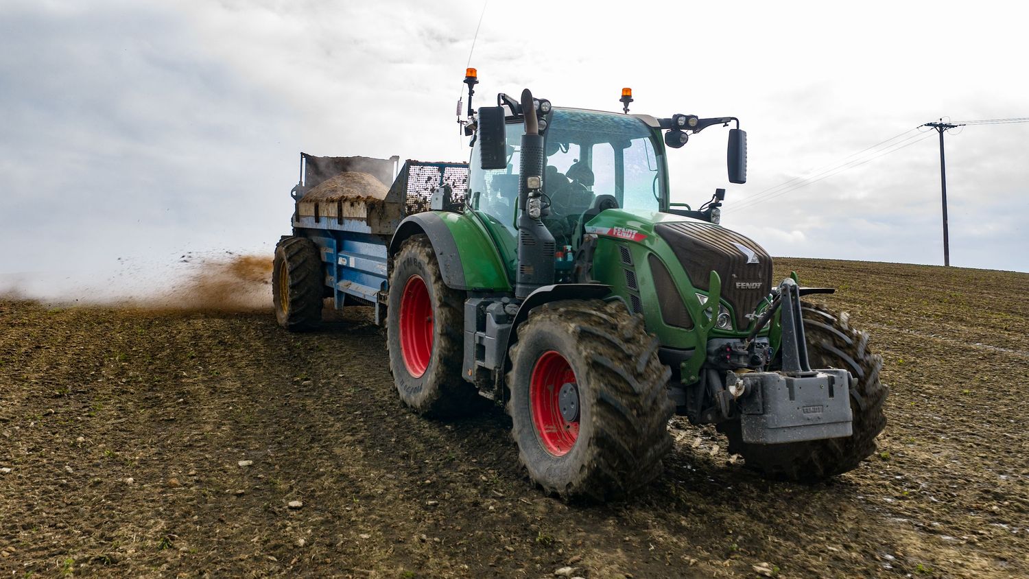 Green Fendt tractor spreading fertilizer on a plowed agricultural field under a cloudy sky.