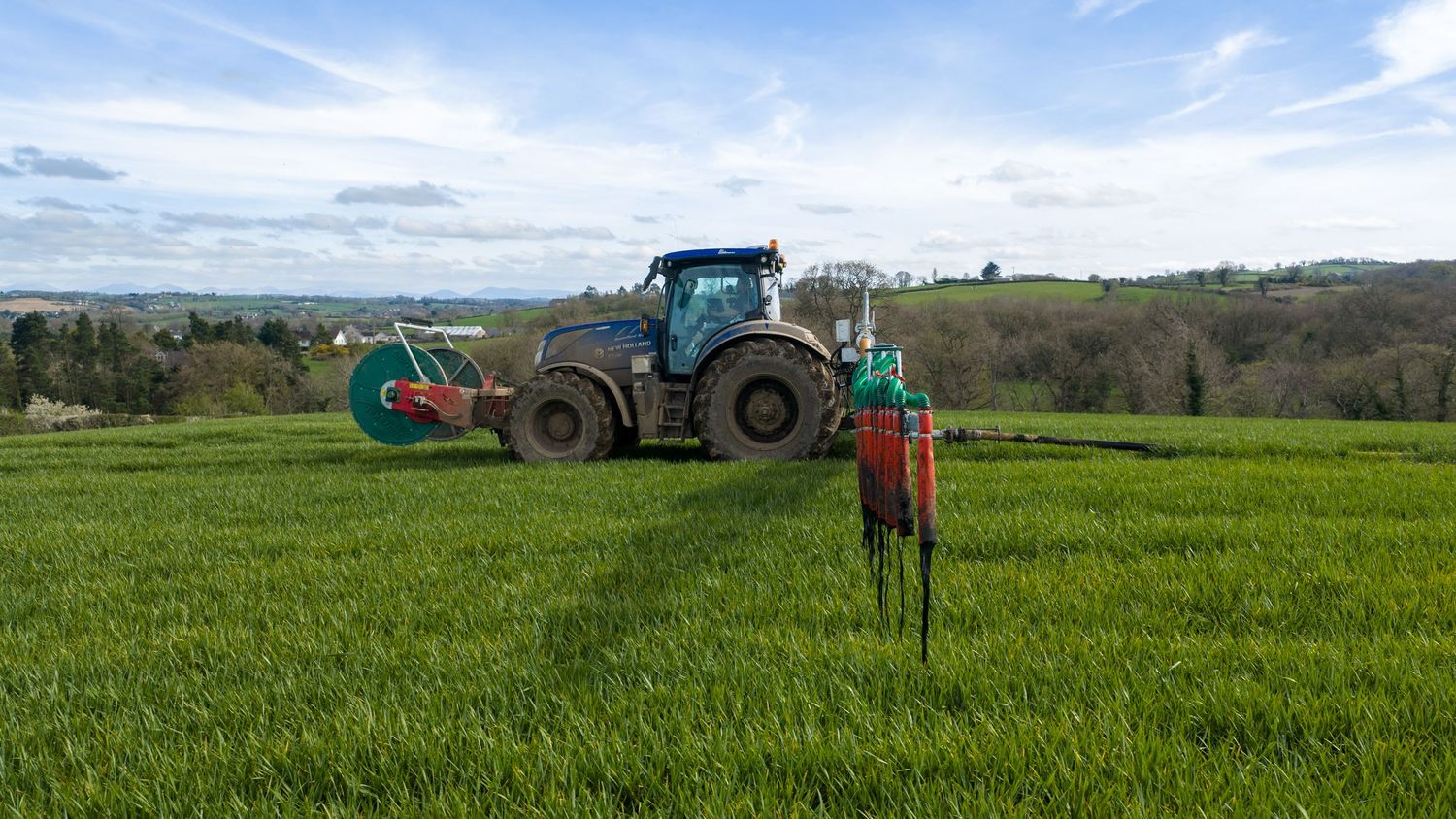 Blue tractor spraying pesticides on lush green crop field with rolling hills and cloudy sky in background.