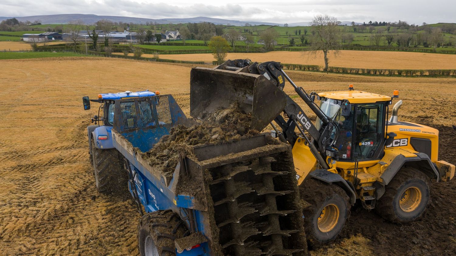 Yellow and blue heavy machinery loaders moving soil in a farm field during agricultural land preparation.