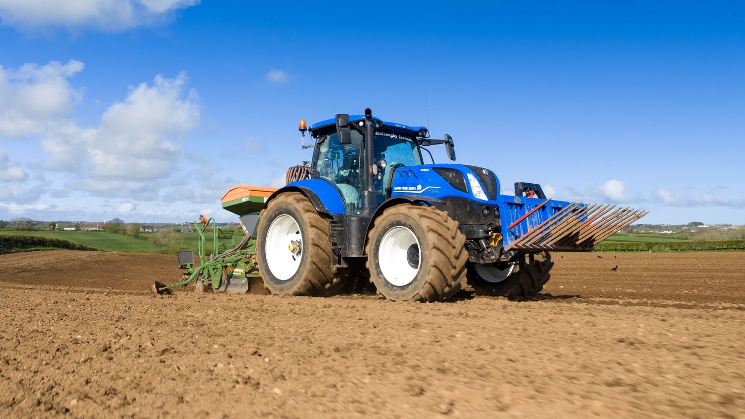 Blue New Holland tractor tilling a brown agricultural field under a partly cloudy sky on a sunny day.