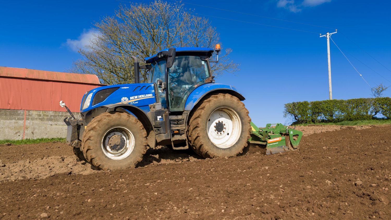 Blue New Holland tractor plowing dark soil in a farm field on a sunny day.