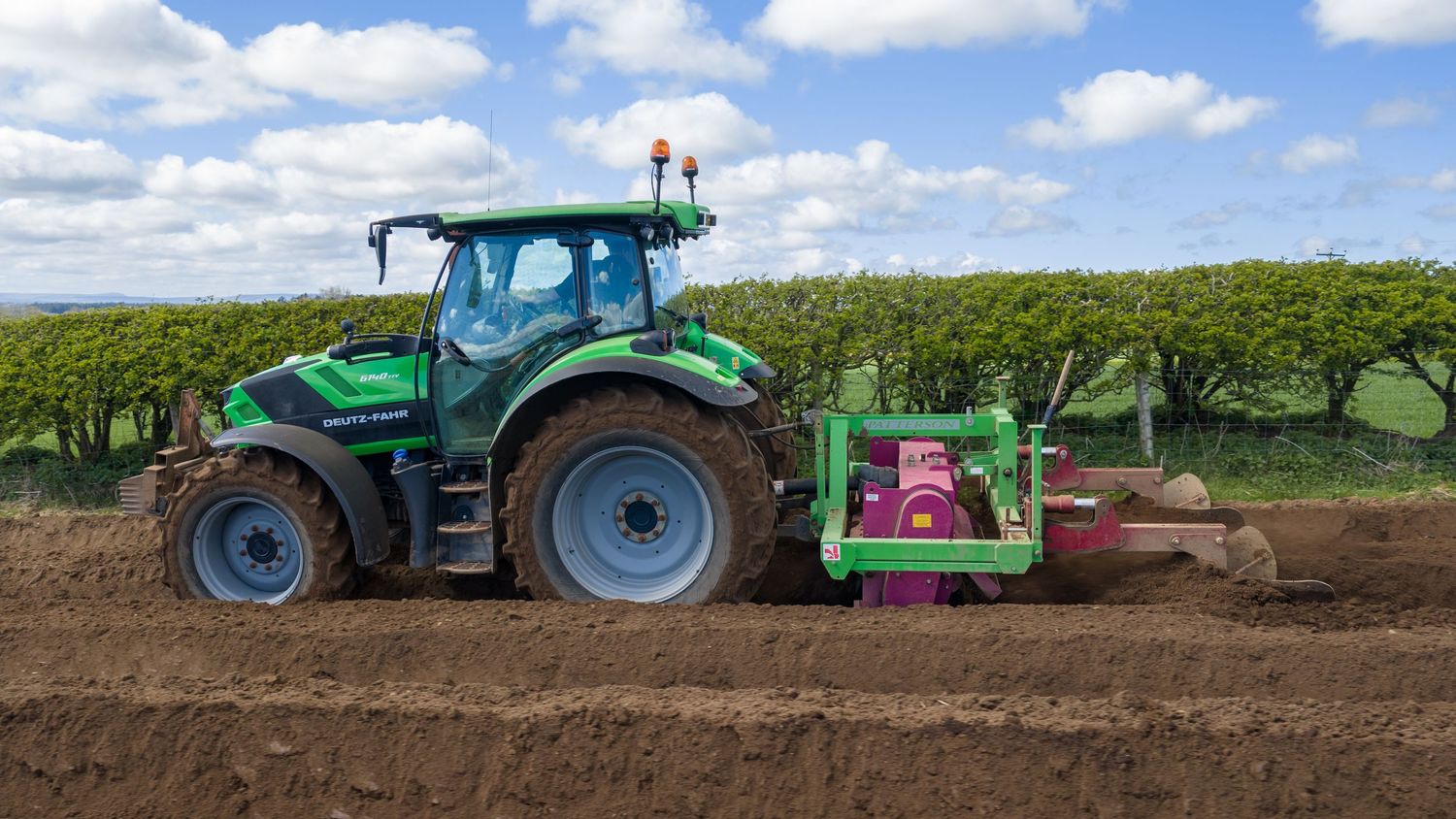 Green tractor with pink attachment tilling dark soil in a farm field under a partly cloudy sky.