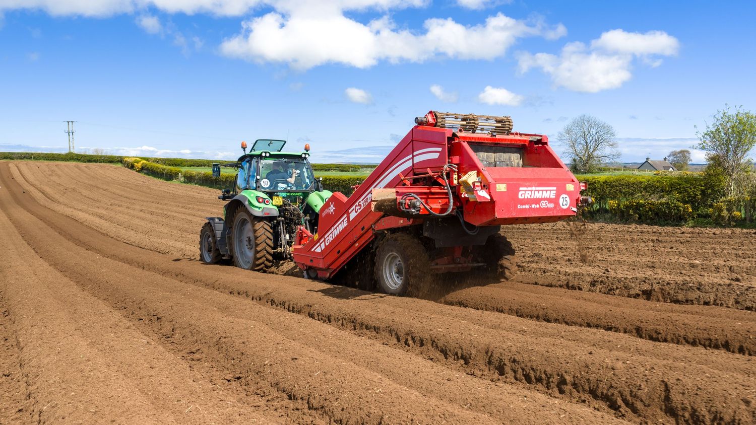 Green tractor pulling a red harvesting machine across a plowed agricultural field on a sunny day.