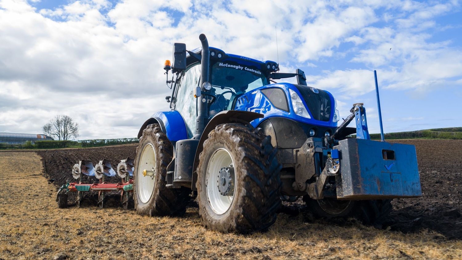 Blue New Holland tractor pulling agricultural equipment across a plowed field under a cloudy sky.