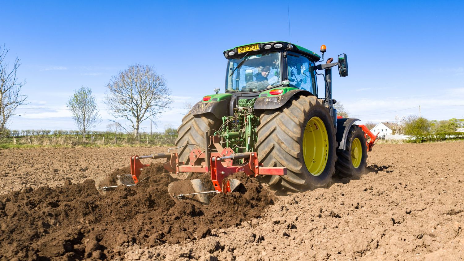 Green John Deere tractor plowing a brown agricultural field under a clear blue sky on a sunny day.