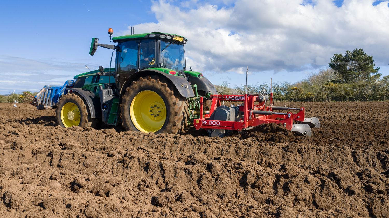 Green John Deere tractor plowing a brown agricultural field with a red tillage implement on a sunny day.