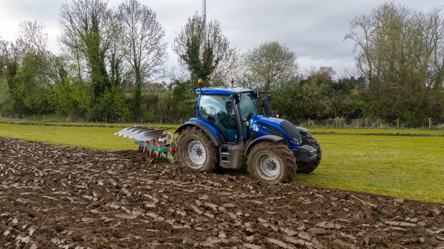 Blue tractor plowing a field with a multi-furrow plow attachment on a cloudy day near green trees.