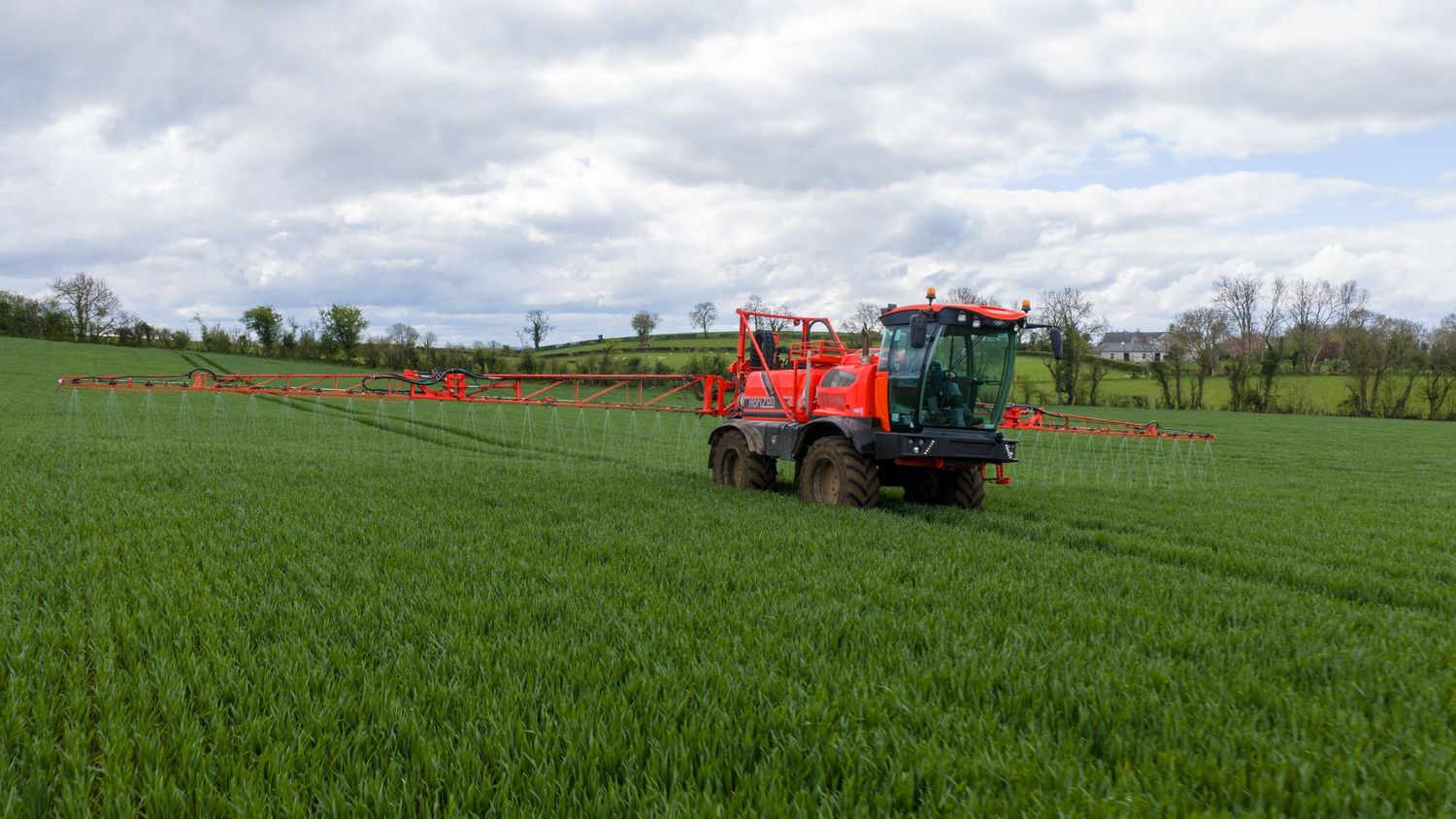 Red agricultural sprayer applying pesticides on lush green wheat field under cloudy sky in rural countryside.