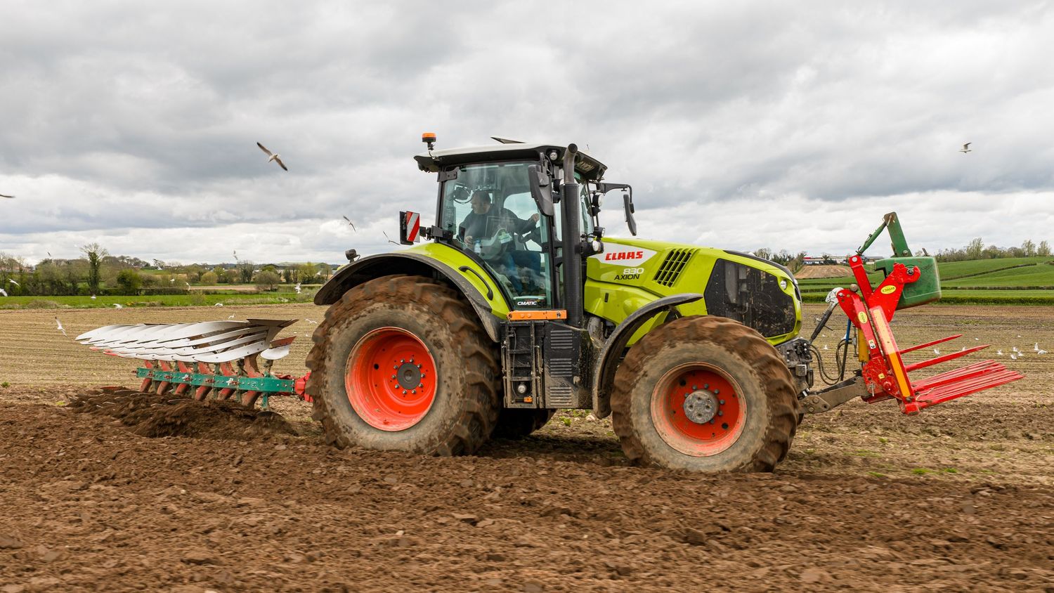 A green CLAAS tractor with red wheels plowing a field under cloudy skies in a rural agricultural setting.