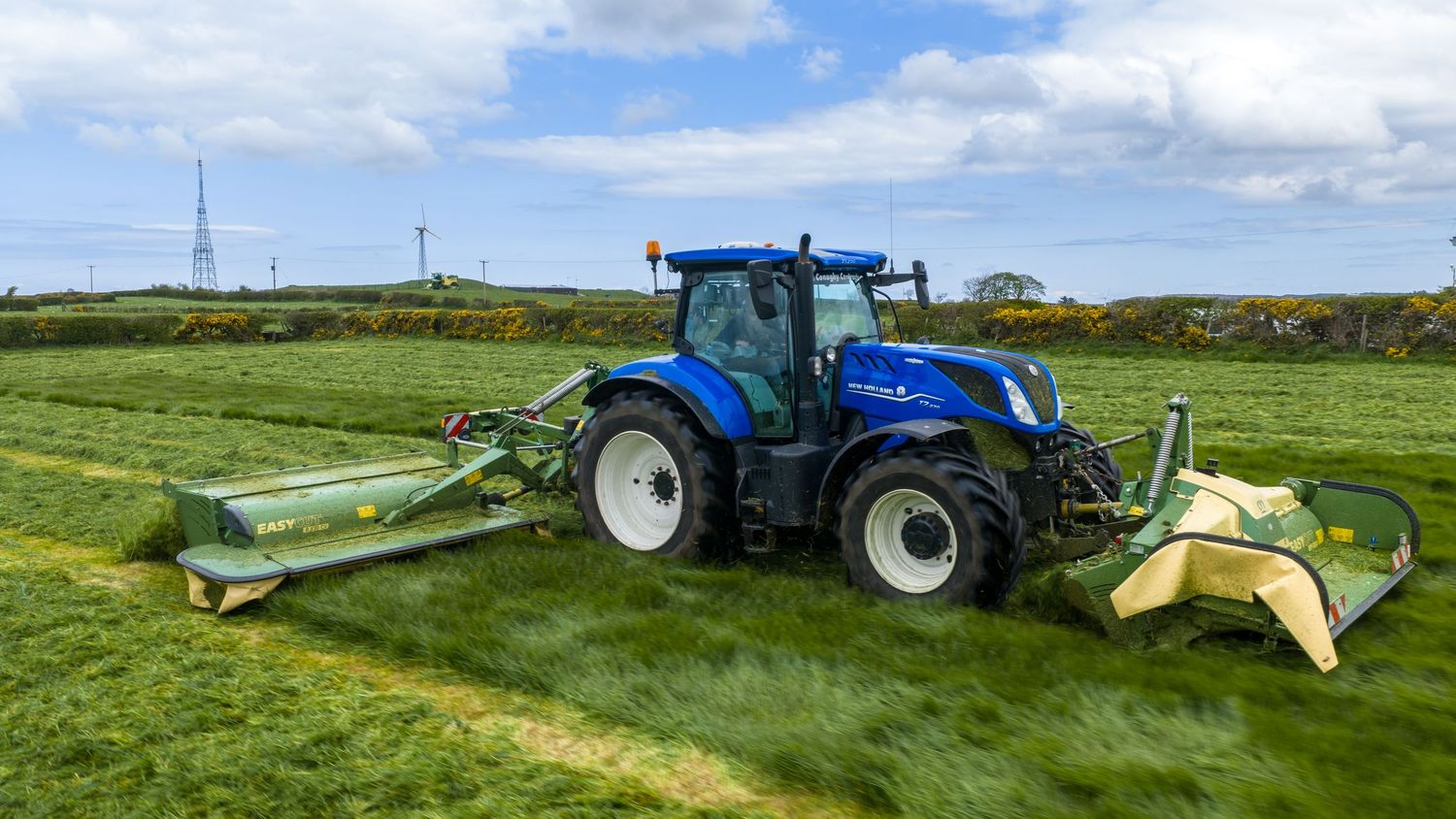 Blue New Holland tractor mowing green grass fields with front and rear mowers attached on a sunny day.