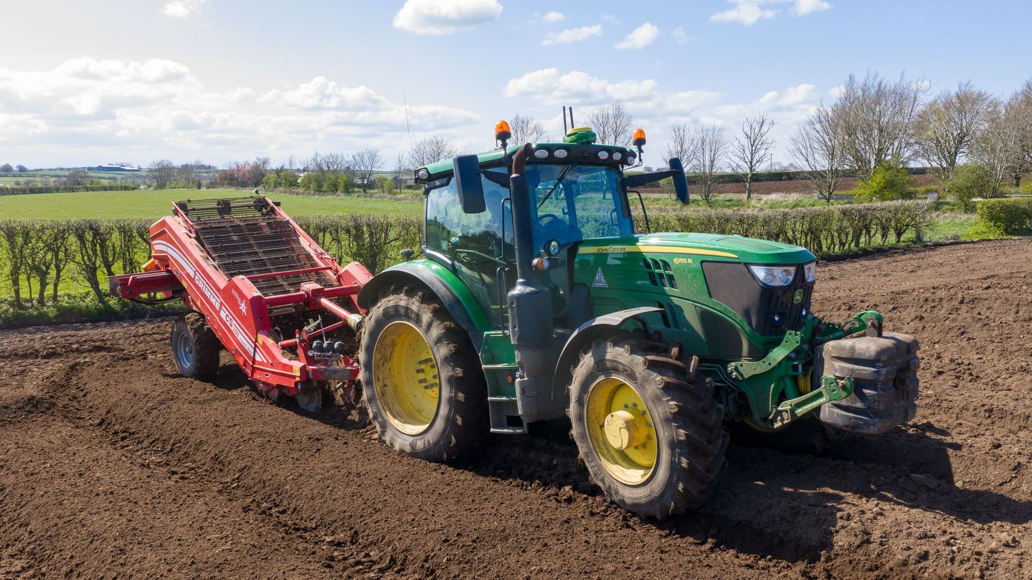 Green John Deere tractor pulling red potato harvester equipment across a freshly plowed agricultural field.