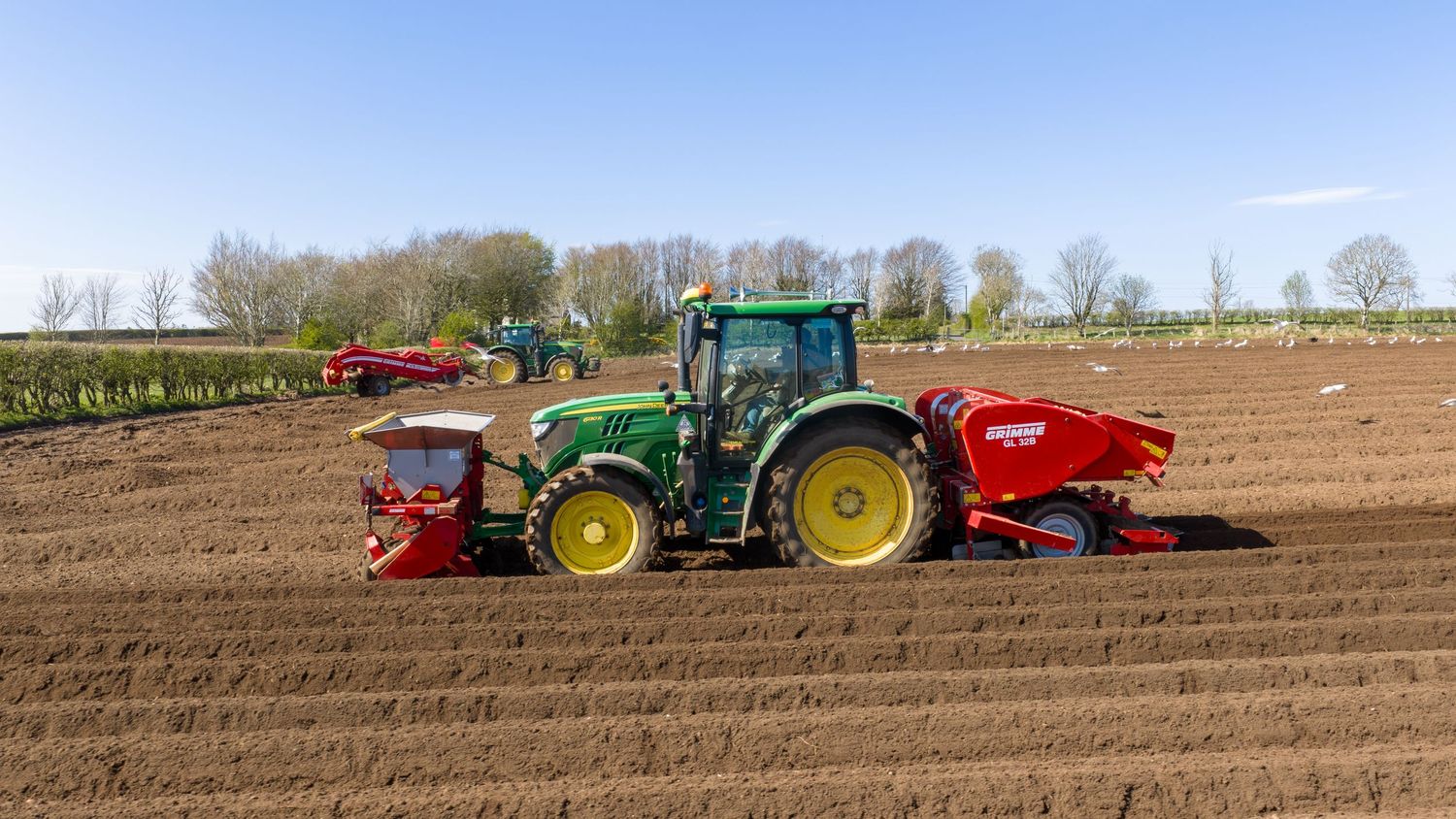 Green John Deere tractor with red planting equipment working in a freshly plowed agricultural field on a sunny day.