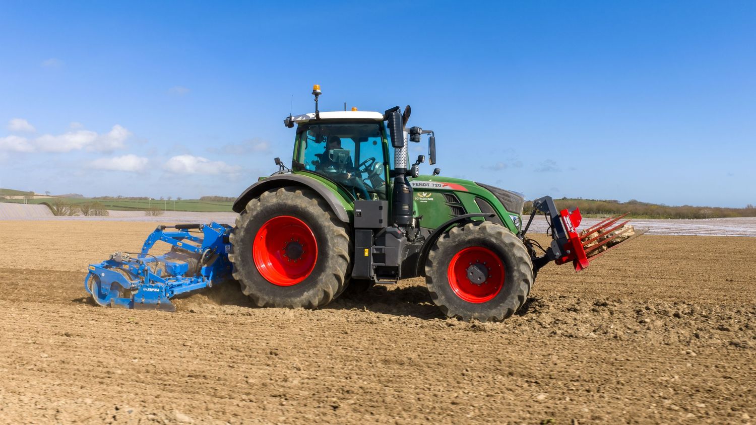 Green tractor with red wheels plowing a dry field under a bright blue sky on a sunny day.