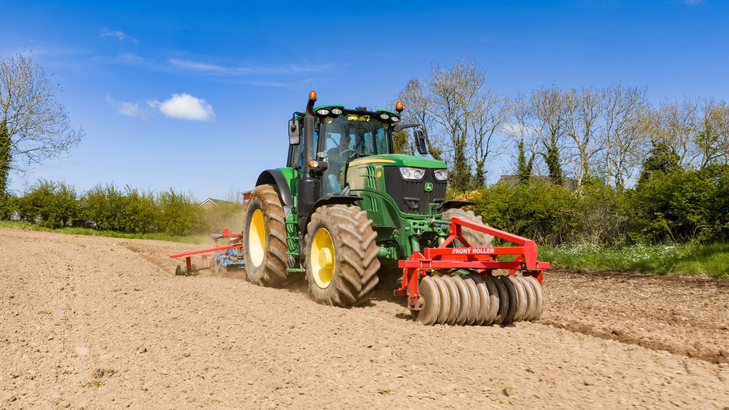Green John Deere tractor plowing a dry field with a red cultivator attachment on a sunny day.