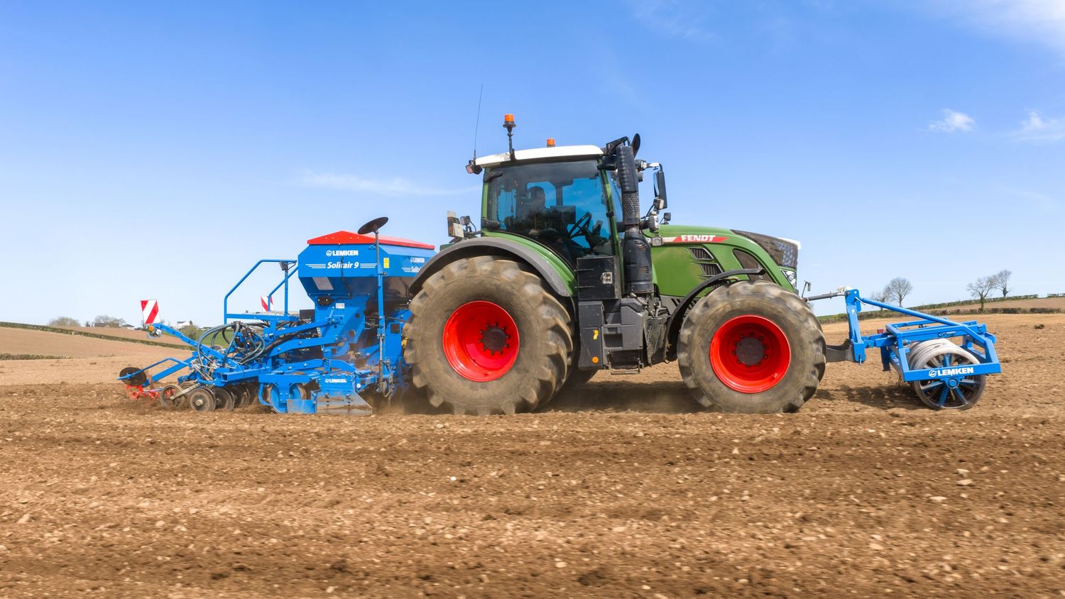 Green tractor with red wheels pulling blue farming equipment across a plowed field under a clear blue sky.