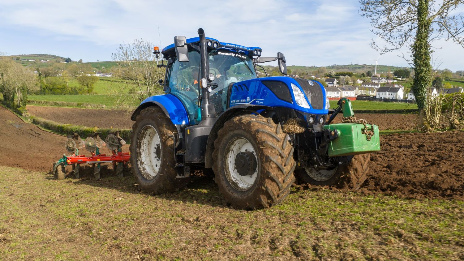 Blue New Holland tractor plowing a field with red cultivator attachment on a cloudy day in rural countryside.