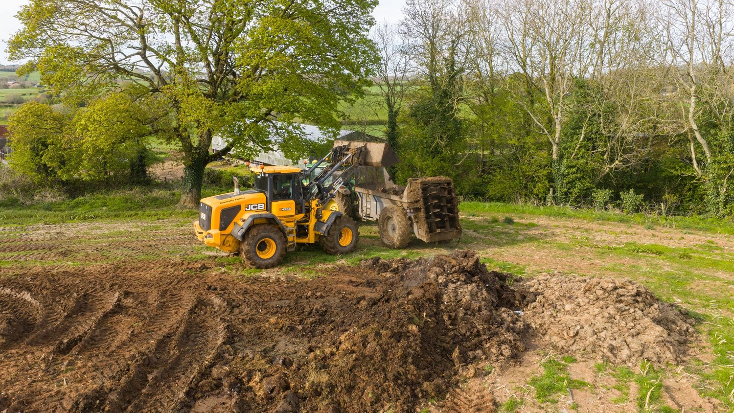 Yellow wheel loader clearing and leveling muddy ground on a rural construction site surrounded by green trees.