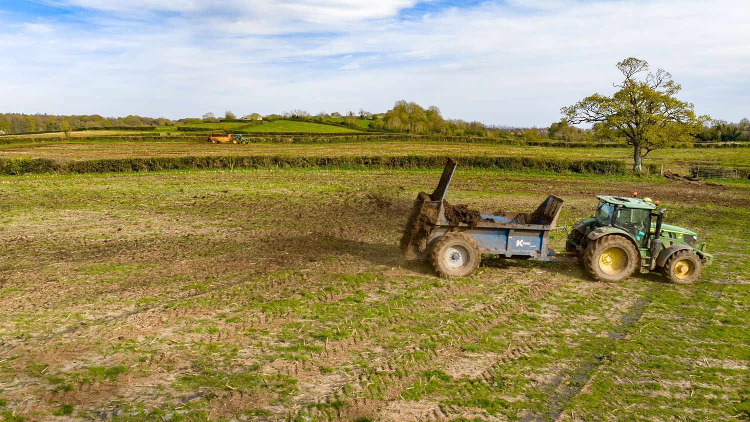 Green tractor spreading manure across a muddy farm field with lush green hedgerows and trees under a blue sky.