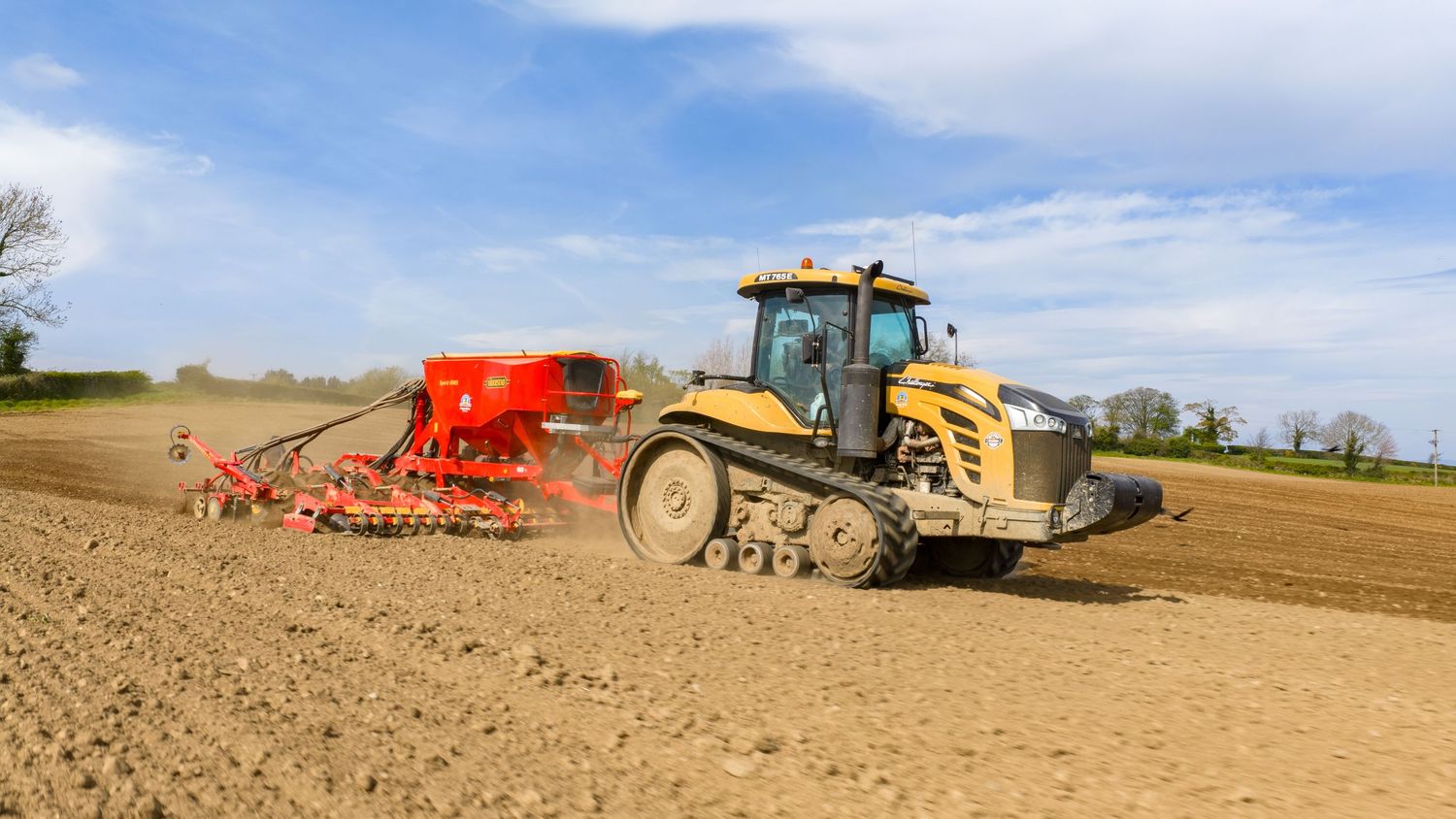 Yellow tracked tractor pulling red seeder equipment across a plowed agricultural field under blue skies.