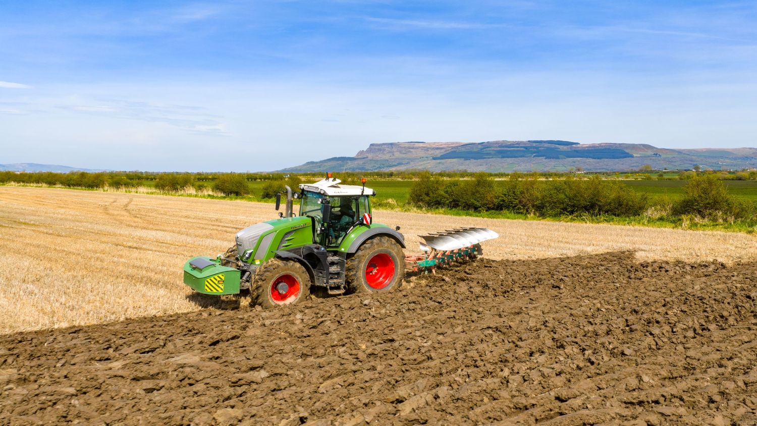 Green Fendt tractor plowing a field with mountains in the background under a blue sky.