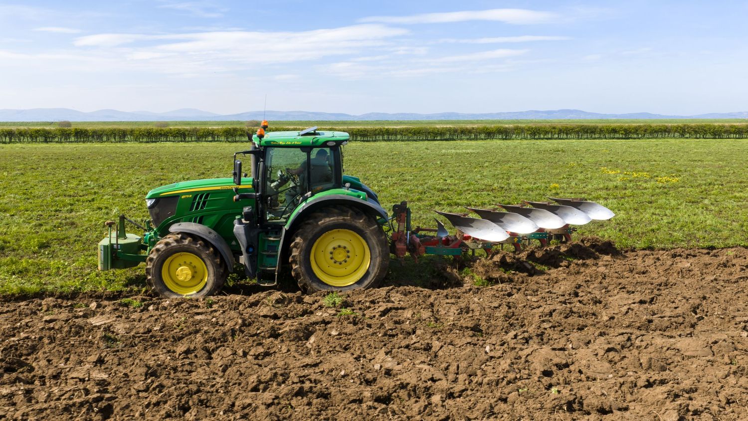 Green John Deere tractor plowing a field with a multi-bottom plow on a sunny day in a rural farm setting.
