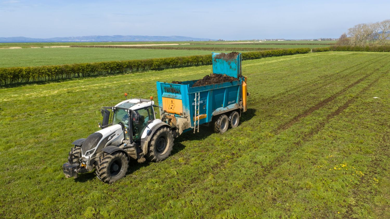 Aerial view of a white tractor spreading manure on green farmland with a blue trailer under clear skies.