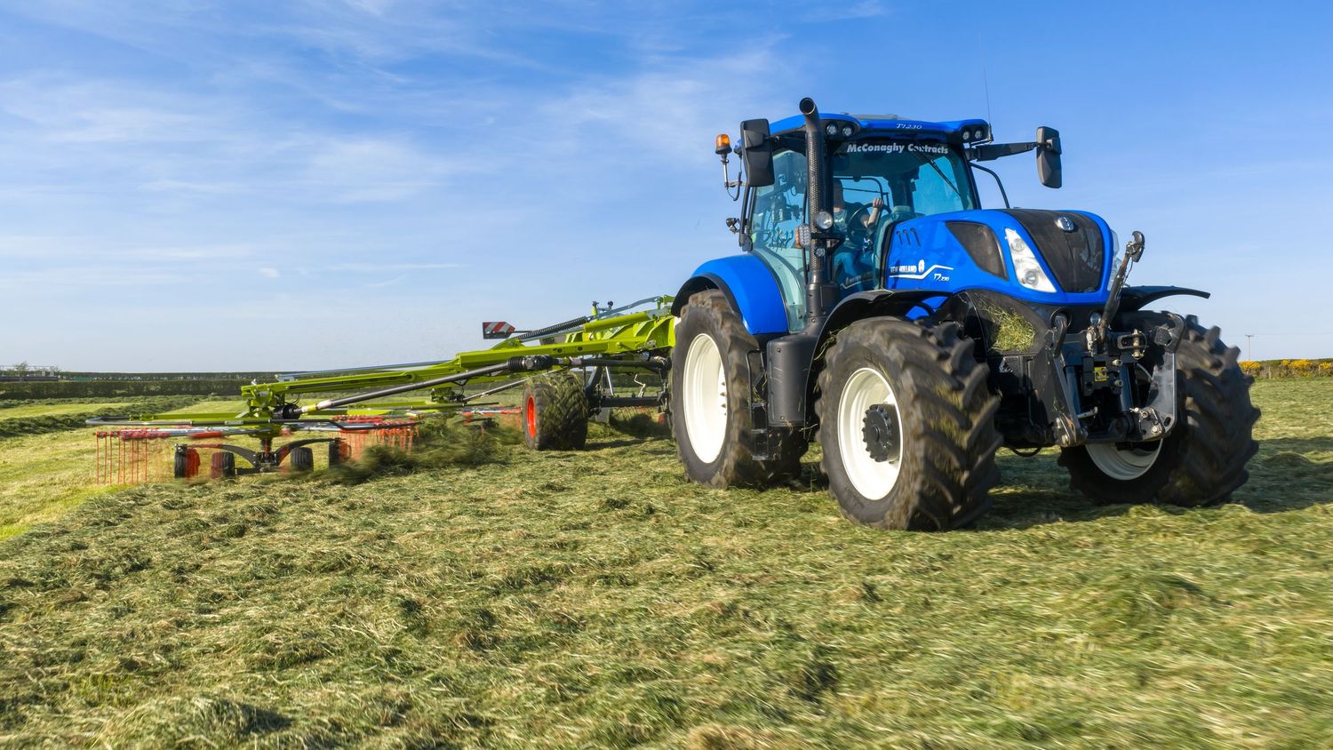 Blue New Holland tractor raking hay in a green field on a sunny day.