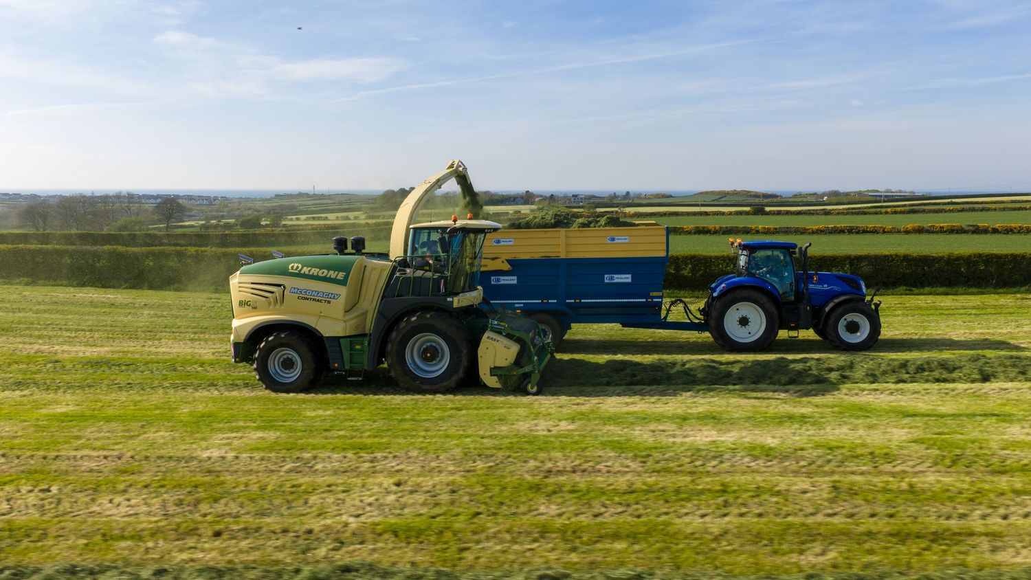 Large green forage harvester and blue tractor working together in lush green fields under a clear blue sky.