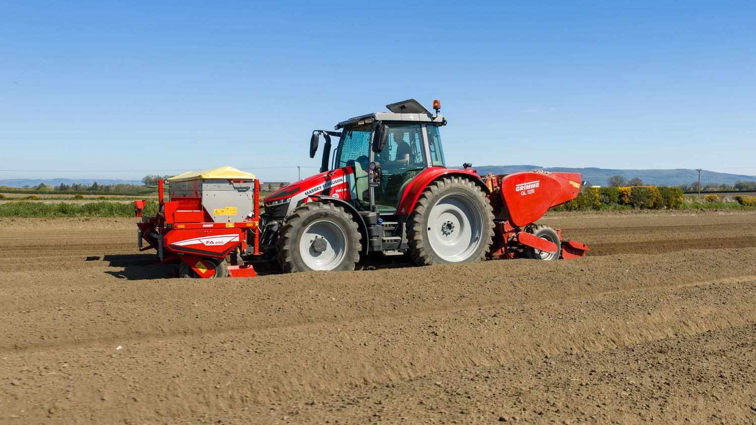 Red tractor with agricultural planting equipment working in a freshly tilled field under a clear blue sky.