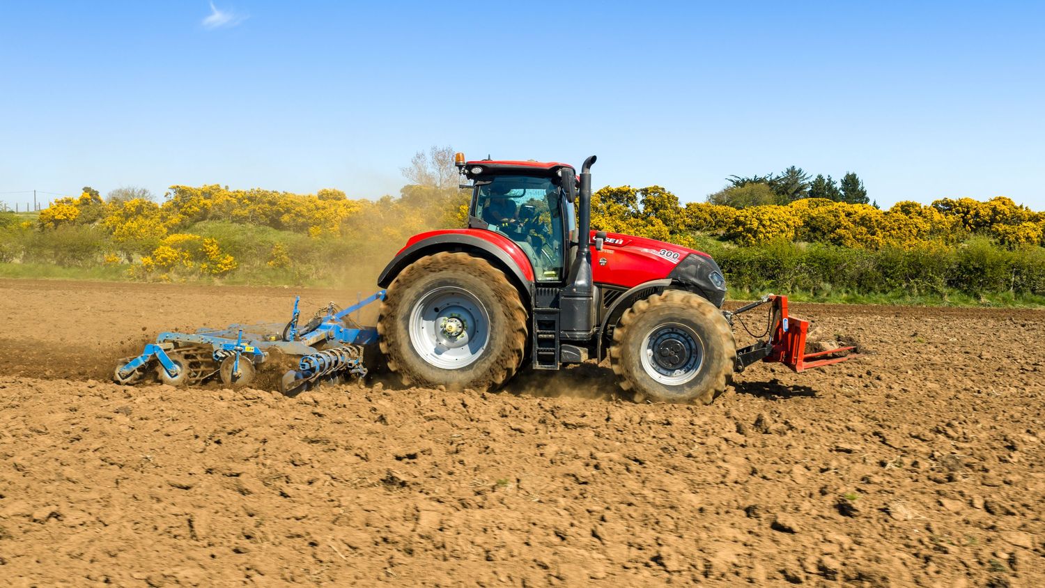 Red tractor plowing a brown agricultural field on a sunny day with yellow flowering shrubs in the background.