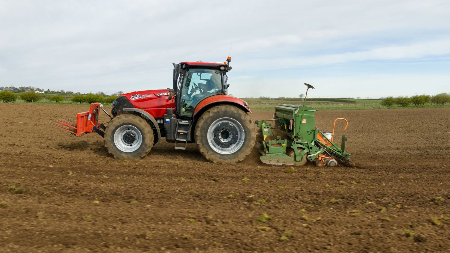 Red Case IH tractor with green planting equipment working in a freshly tilled agricultural field on a cloudy day.