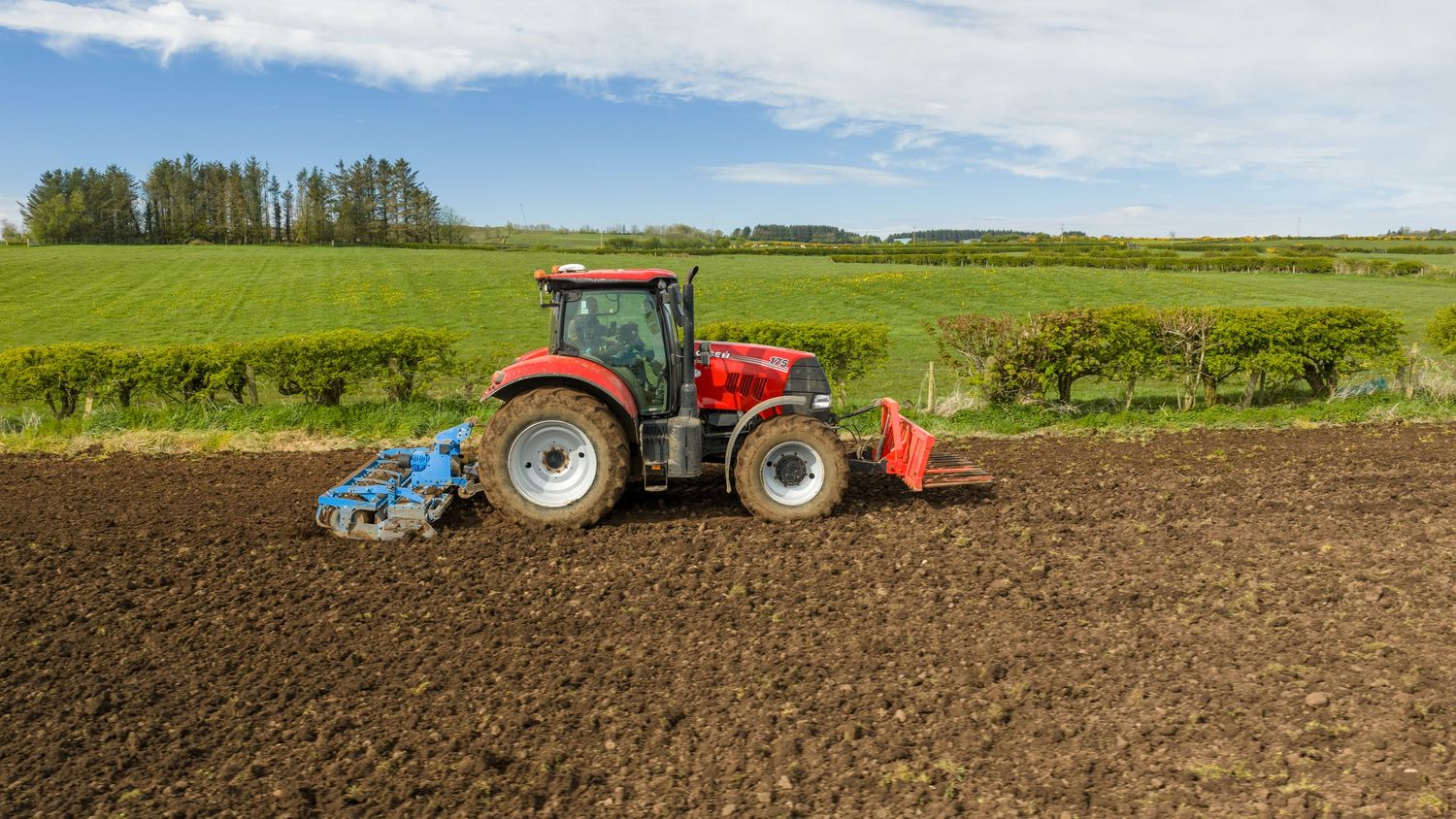 Red tractor plowing a fertile green field under a bright blue sky on a sunny day.