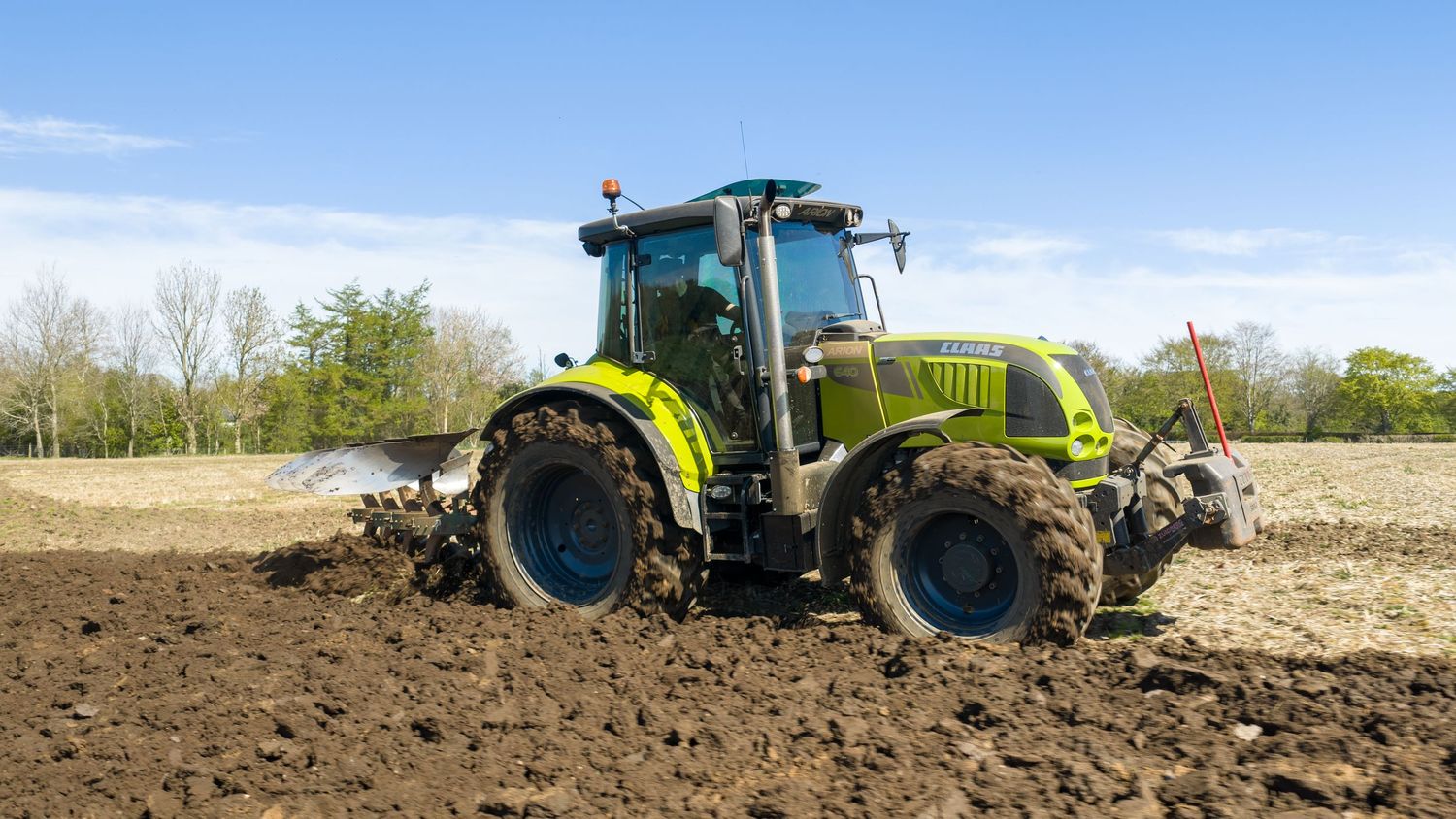 Green tractor plowing dark soil in an open field under a bright blue sky with trees in the background.