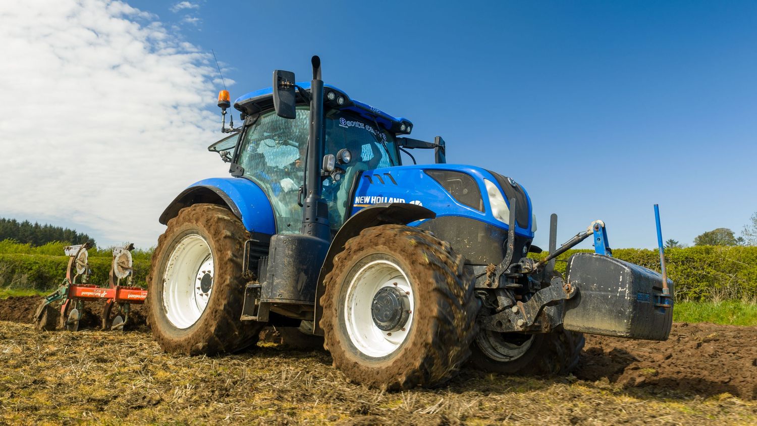 Blue New Holland tractor plowing a field on a sunny day with green hills in the background.