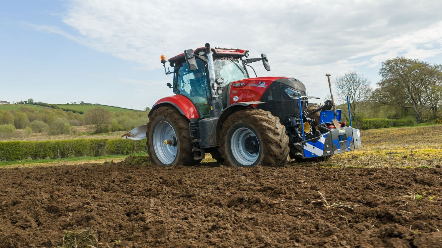 Red Case IH tractor plowing a dark soil field with a blue implement attached, on a sunny day in a green rural landscape.