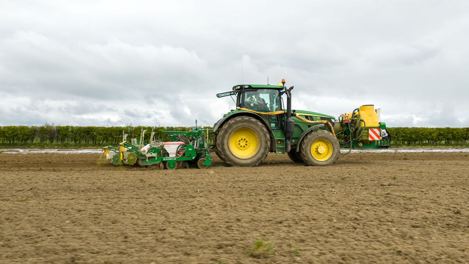 Green John Deere tractor with planting equipment working in a large agricultural field under cloudy skies.