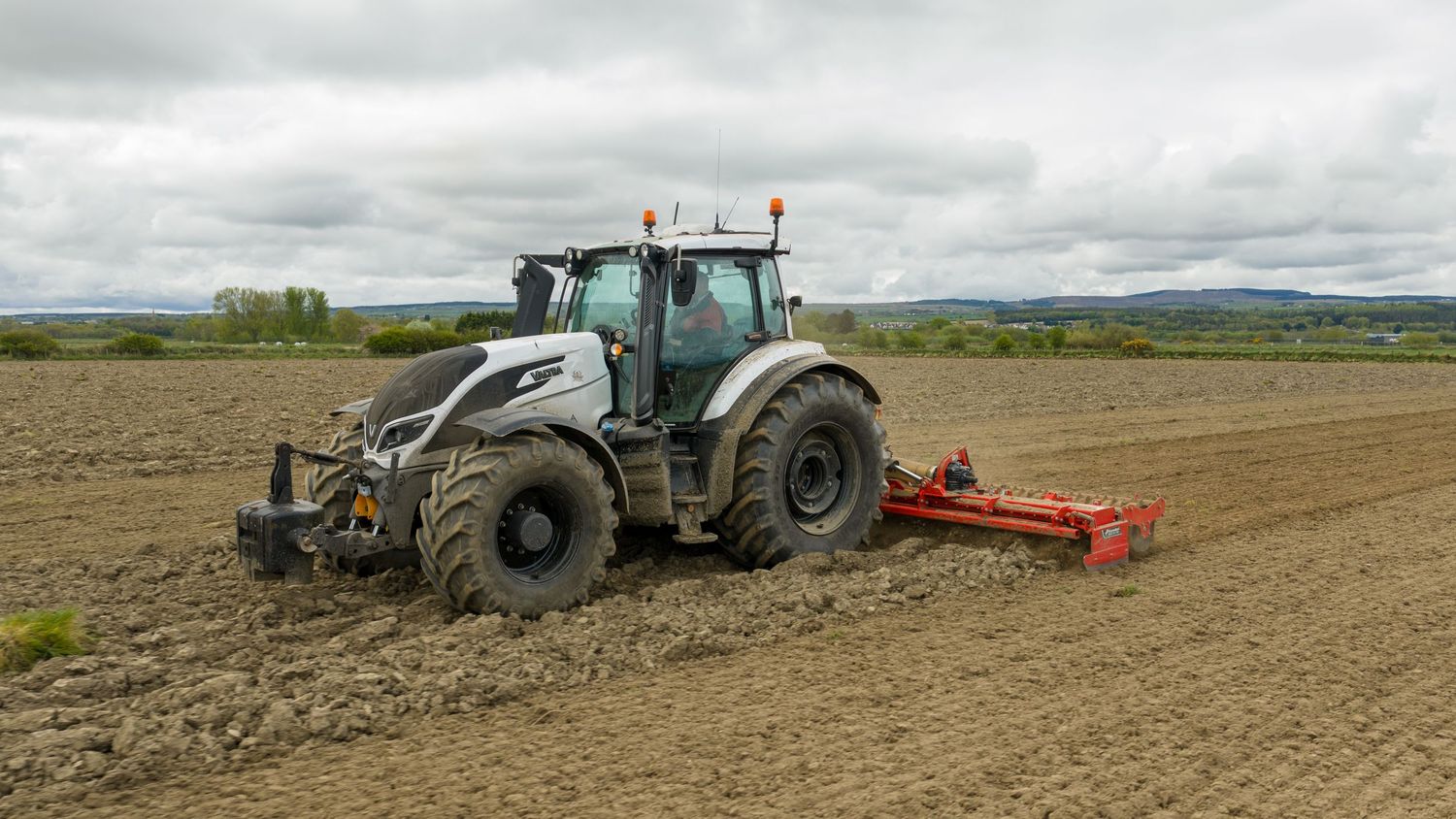 White tractor plowing a large agricultural field under cloudy skies, preparing soil for planting with a red tillage implement.