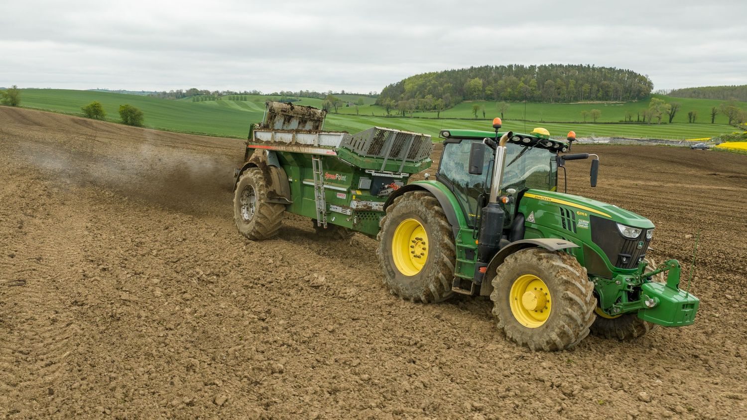 Green John Deere tractor pulling agricultural equipment across a freshly plowed brown field on a cloudy day.