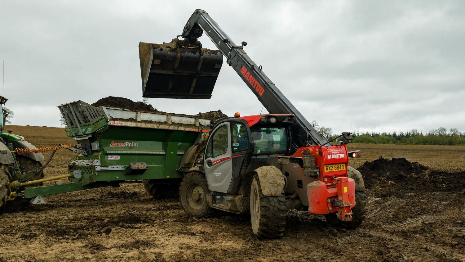 A telehandler loads dirt into a green dump trailer on a muddy construction site with overcast skies.