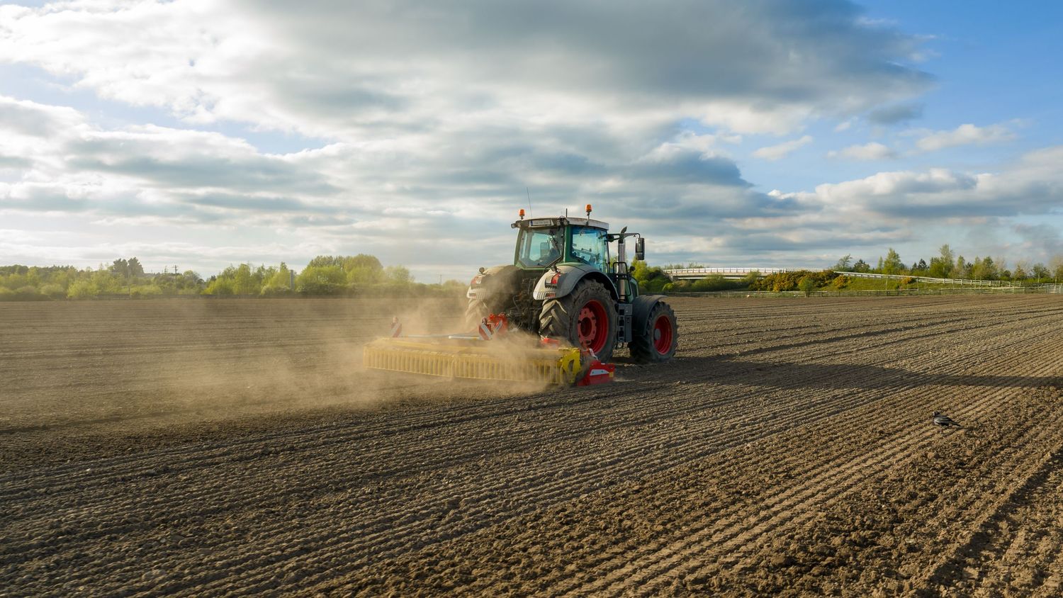 Green tractor plowing dusty farmland field at sunset with dramatic cloudy sky in background.