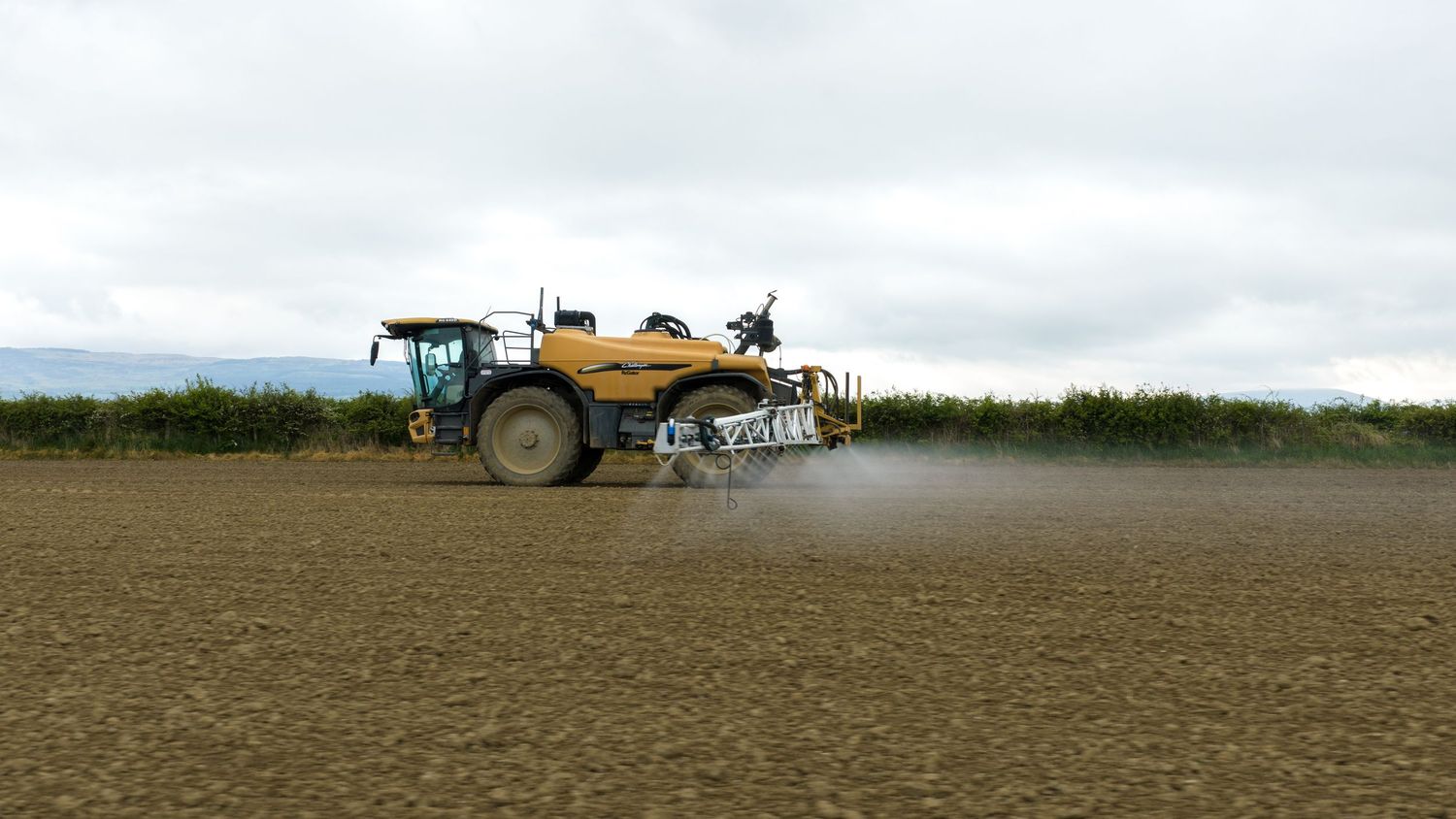 Yellow agricultural sprayer applying pesticides on a large plowed field under cloudy skies.
