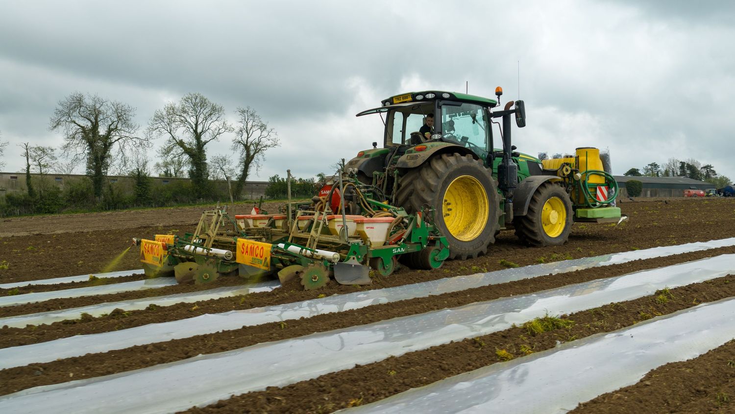 John Deere tractor planting crops in a field with white mulch film rows on a cloudy day.