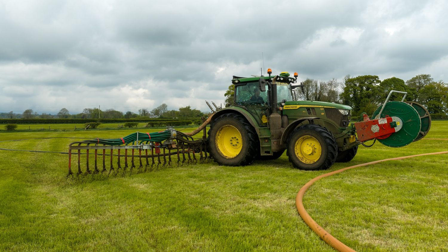 Green John Deere tractor spreading slurry on lush farmland using trailing hose equipment on a cloudy day.