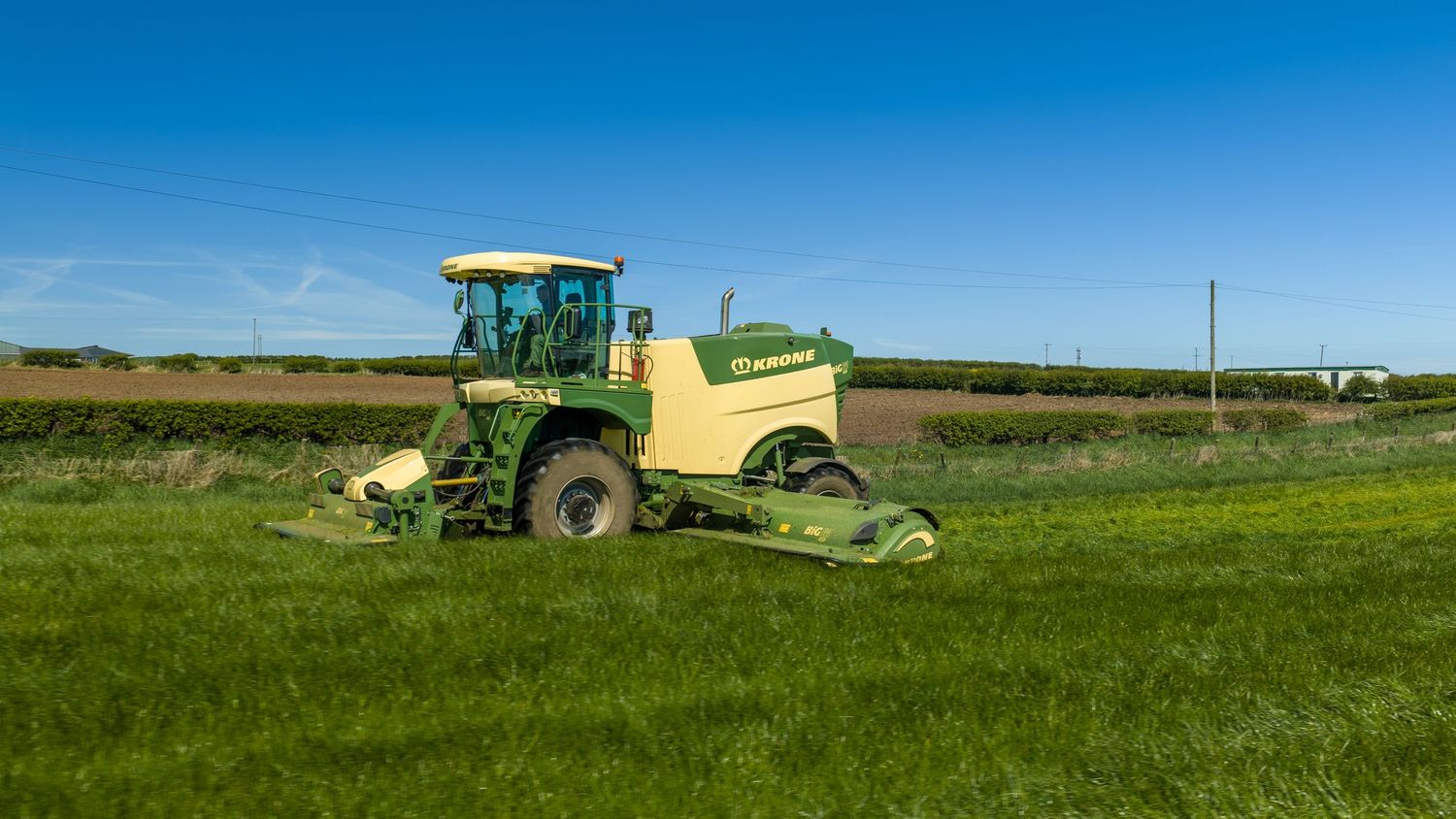 Green Krone self-propelled mower cutting lush grass field under clear blue sky on sunny day.