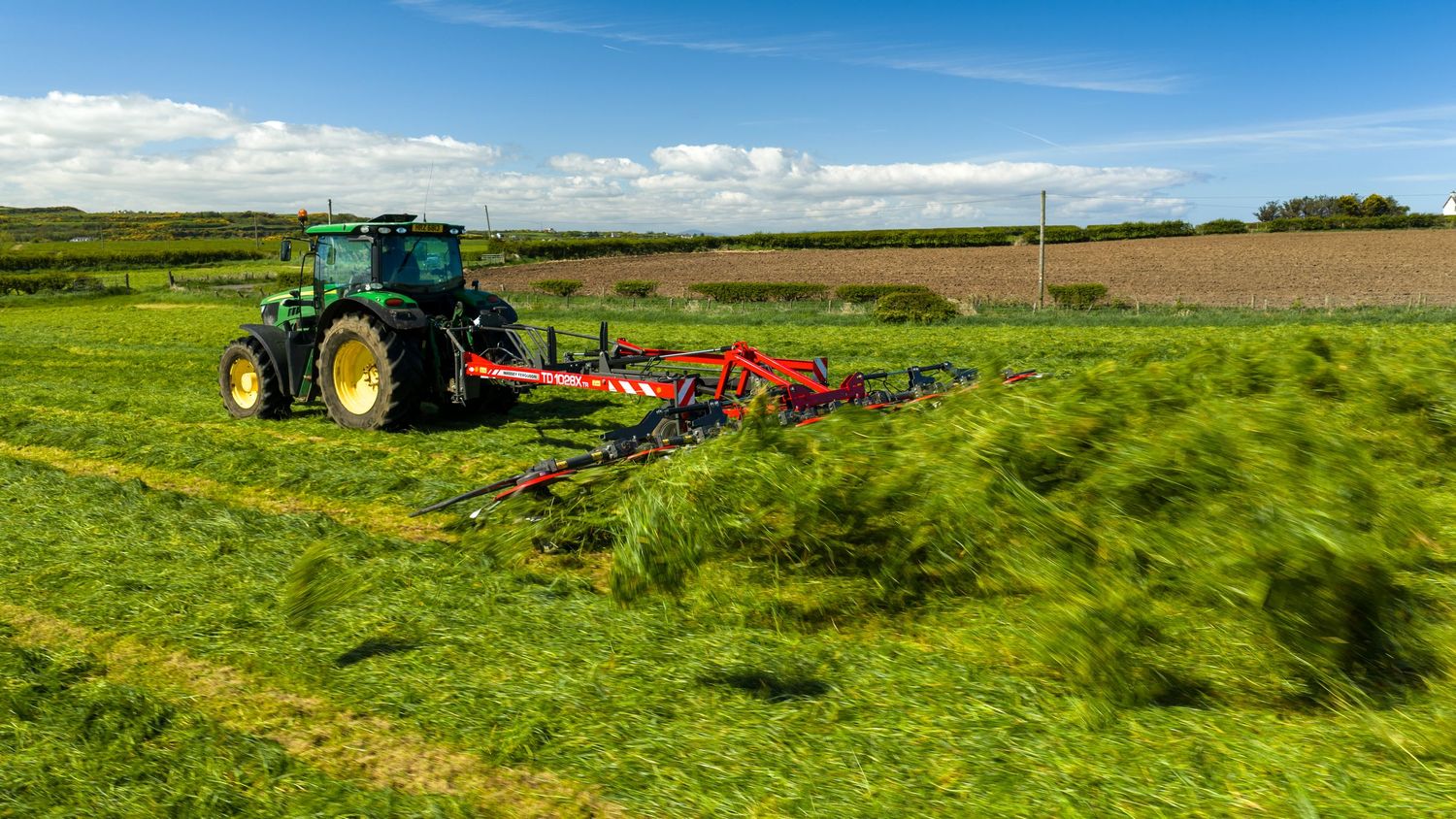 Green tractor pulling red hay rake across lush farm field under blue sky with white clouds.