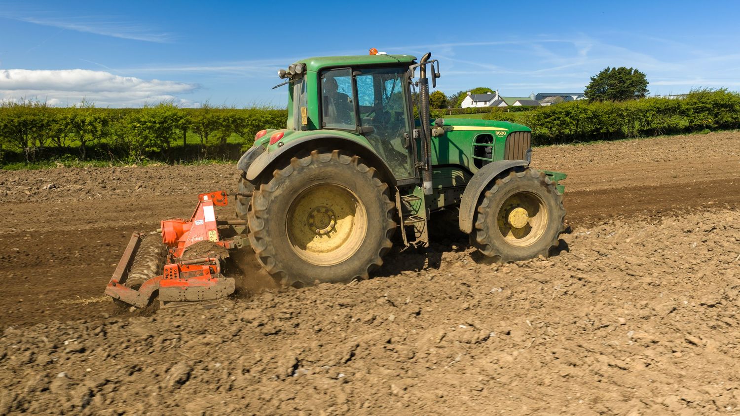 Green John Deere tractor plowing a field on a sunny day with a red attachment tool on fertile farmland.