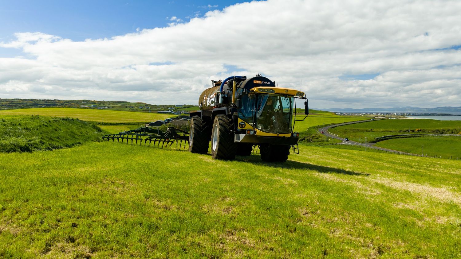 Large blue self-propelled sprayer applying treatment to green agricultural field with coastal views.