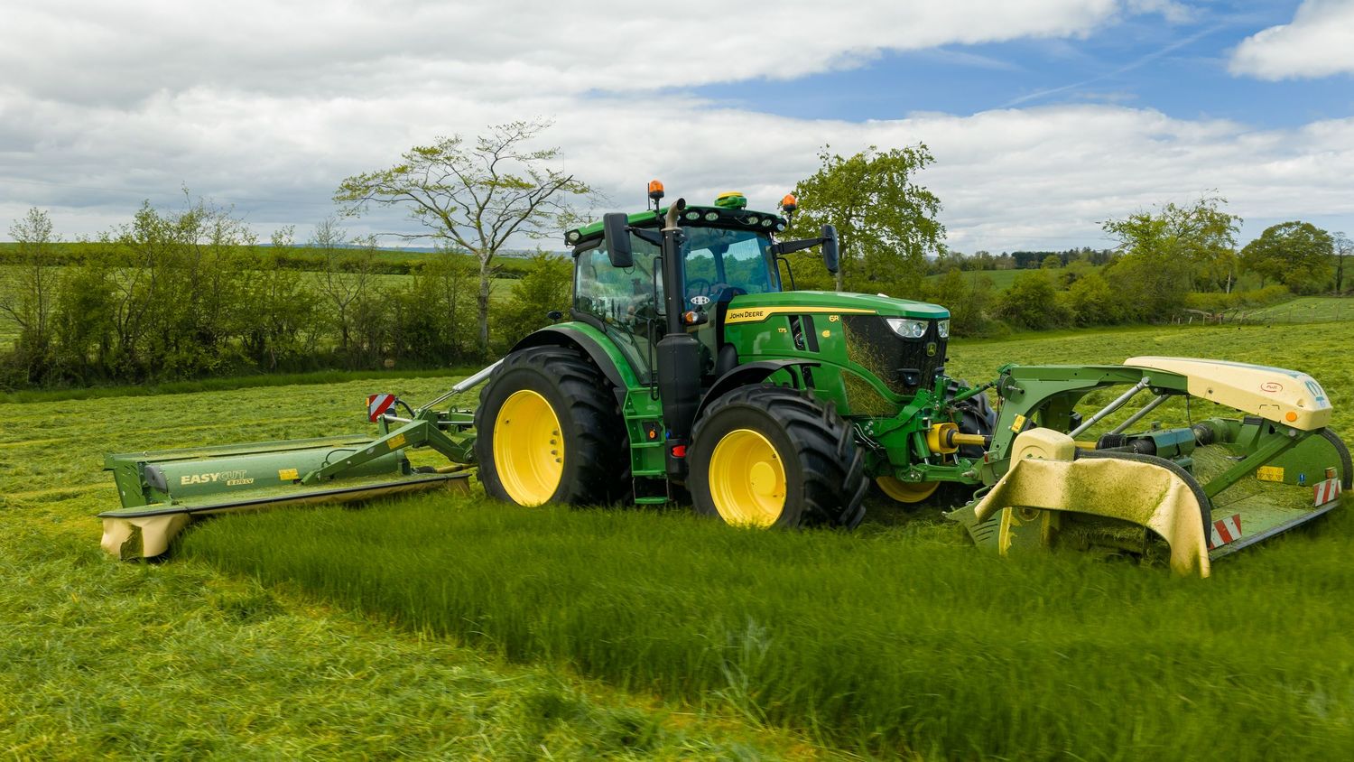 Green John Deere tractor mowing a lush green field with wide cutting attachments on a sunny day.