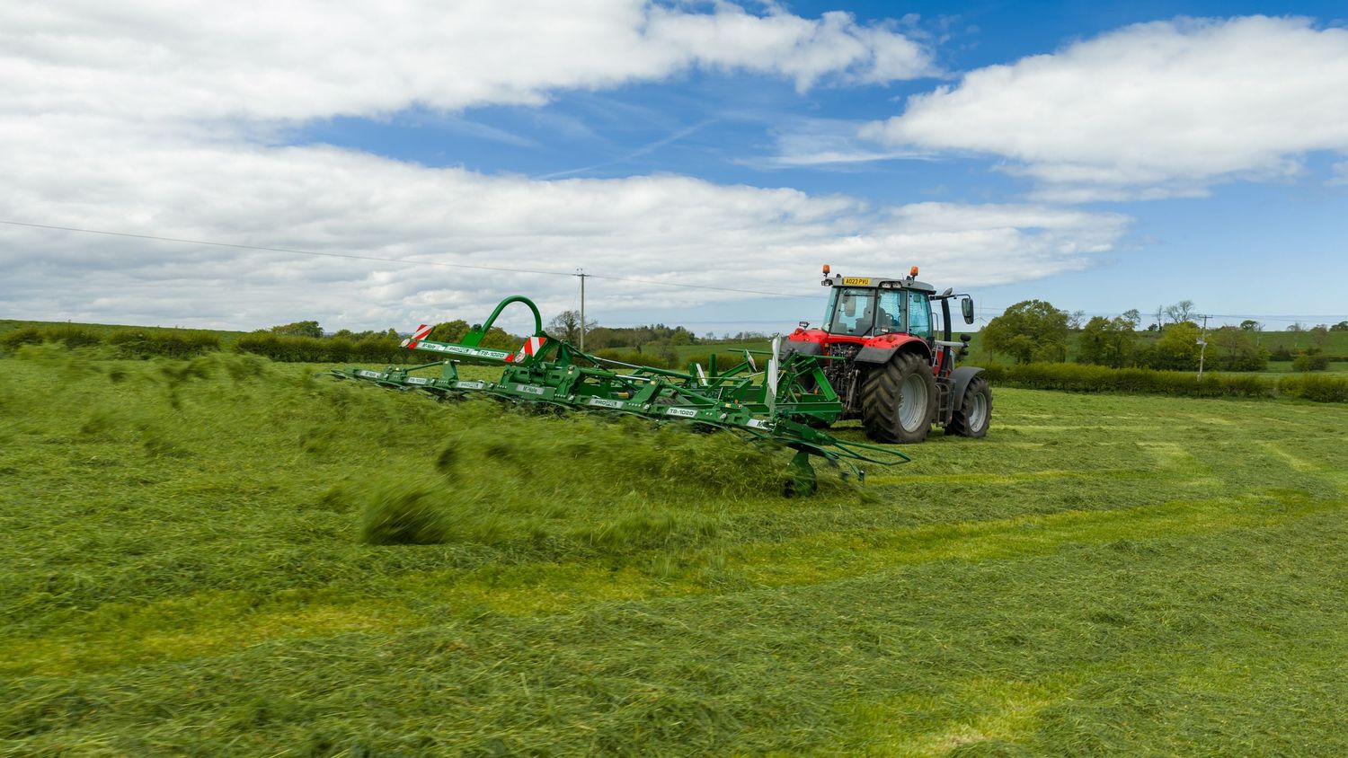 Red tractor pulling a large green hay rake across a lush green field under a partly cloudy sky.